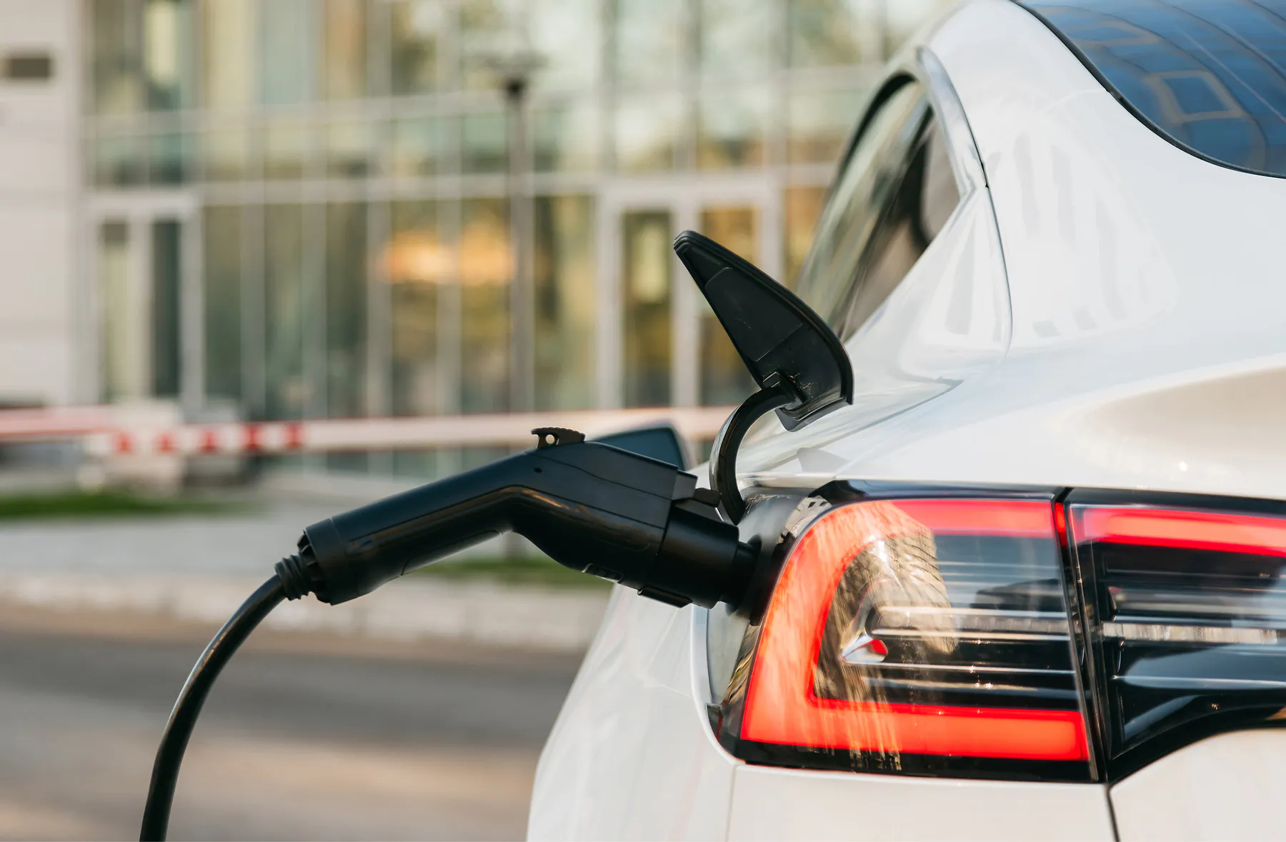 A white electric car is parked and charging at a charging station, with the charging cable plugged into the rear of the vehicle near the taillight. A modern building is visible in the background.