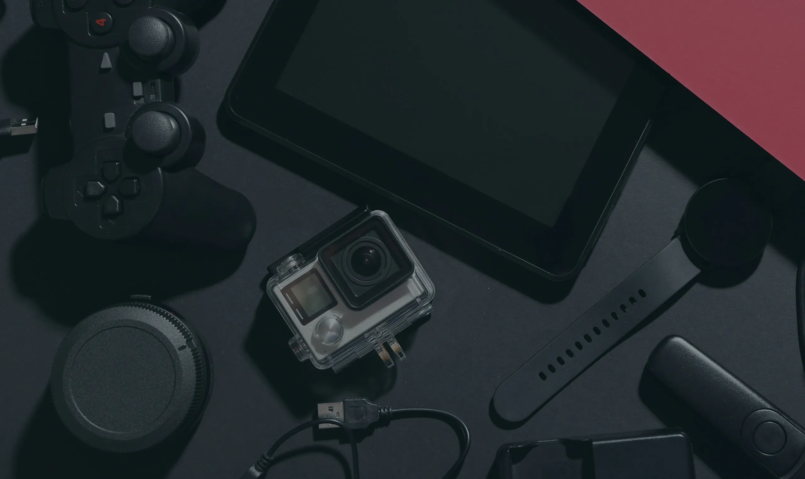 A flat lay of various black electronic gadgets on a dark surface, including a tablet, action camera, smart watch, game controllers, a portable speaker, and charging cables.